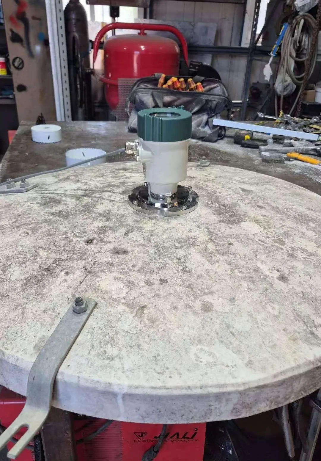 An 80 GHz radar level sensor bolted through a stainless flange in the centre of a circular concrete silo lid, assembled on a workbench