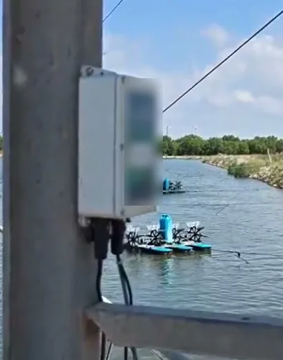 Omni Genesis controller mounted on a concrete post at the pond bank, cable running down toward the water and an aerator floating behind